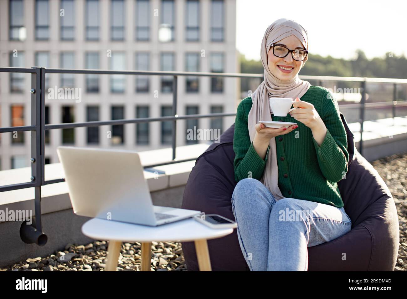 Portrait of young female in hijab drinking coffee while placing laptop ...