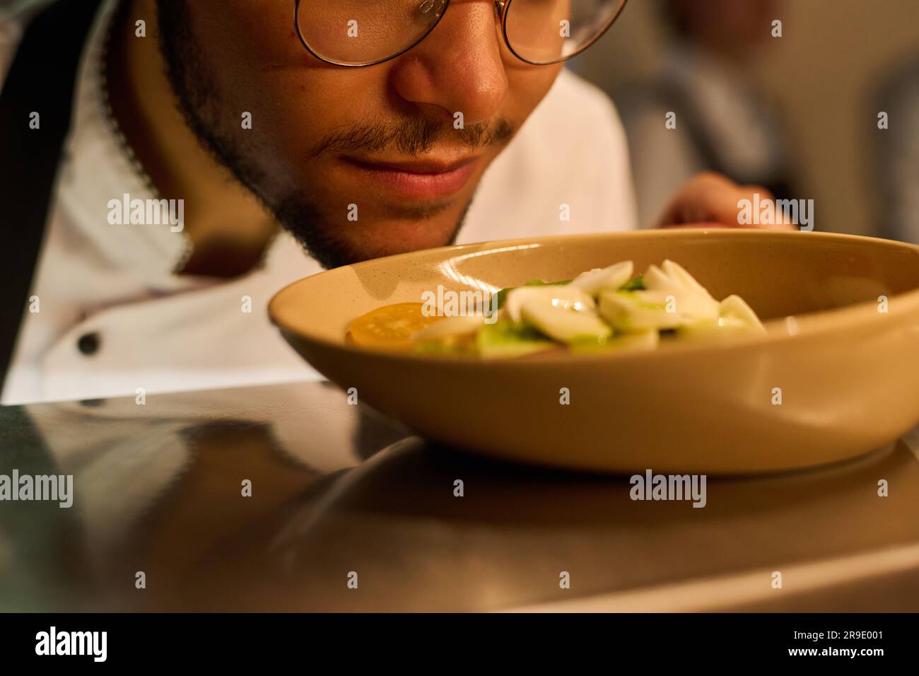 Close-up of face of young male chef bending over plate with fresh vegetable salad with mozarella cheese and sauce while smelling it Stock Photo