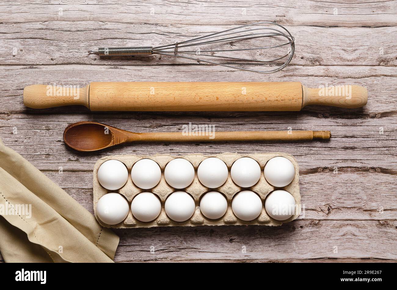 A whisk, a rolling pin, a wooden spoon, some white eggs in an egg container and a piece of cloth on a wooden table. Stock Photo