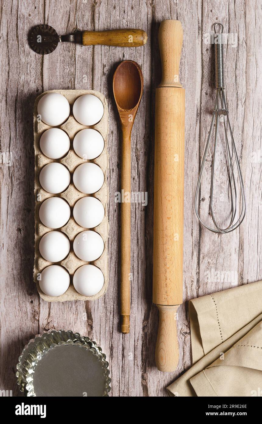 A whisk, a rolling pin, a wooden spoon, some white eggs in an egg container, some little pans, a pasta cutter and a piece of cloth on a wooden table. Stock Photo