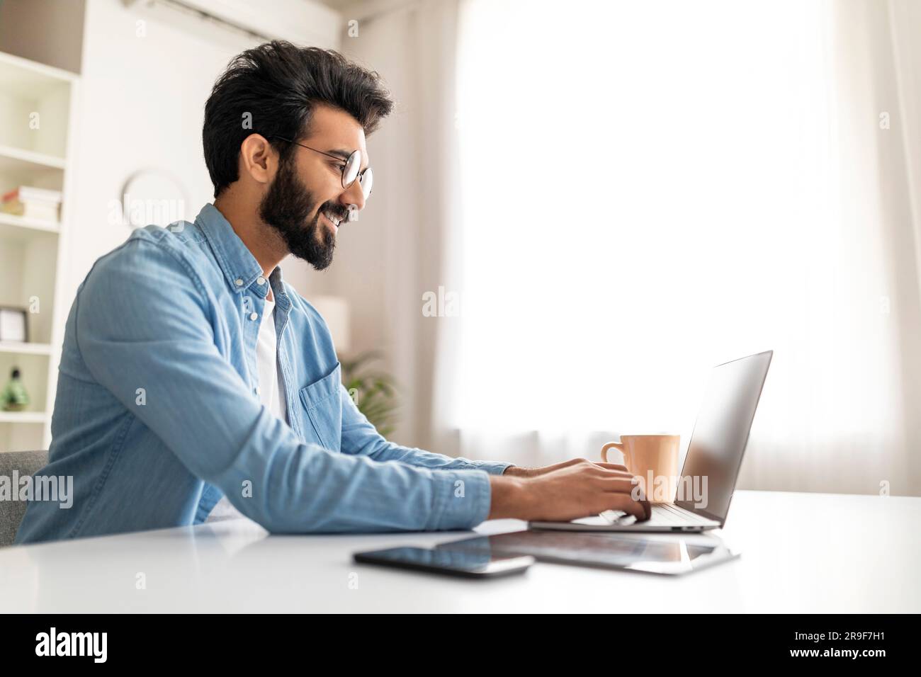 Smiling Indian Male Writer Using Laptop While Working At Home Office Stock Photo
