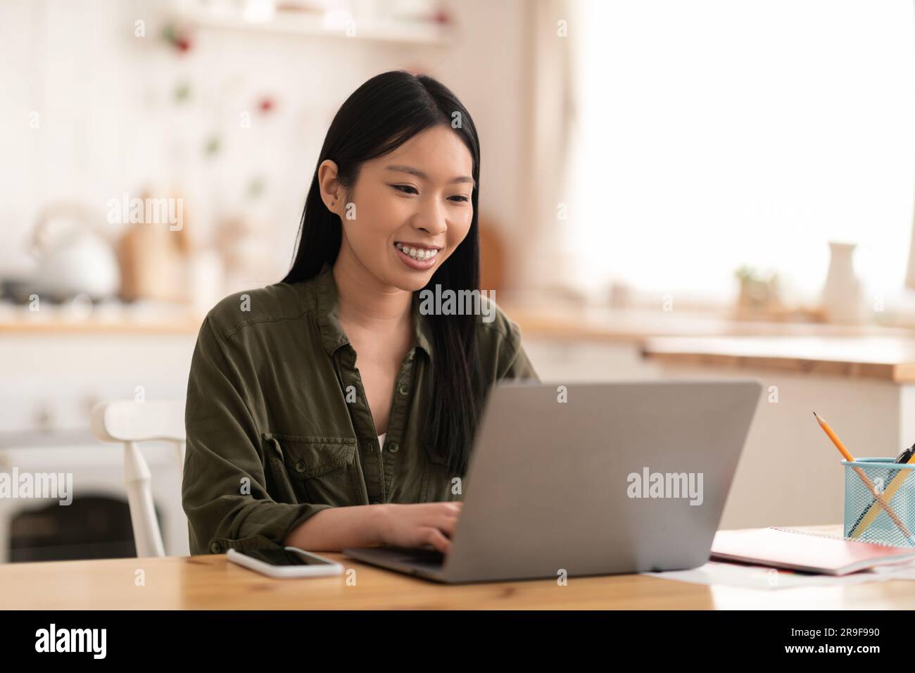 Smiling young asian woman sitting at kitchen, using laptop Stock Photo ...