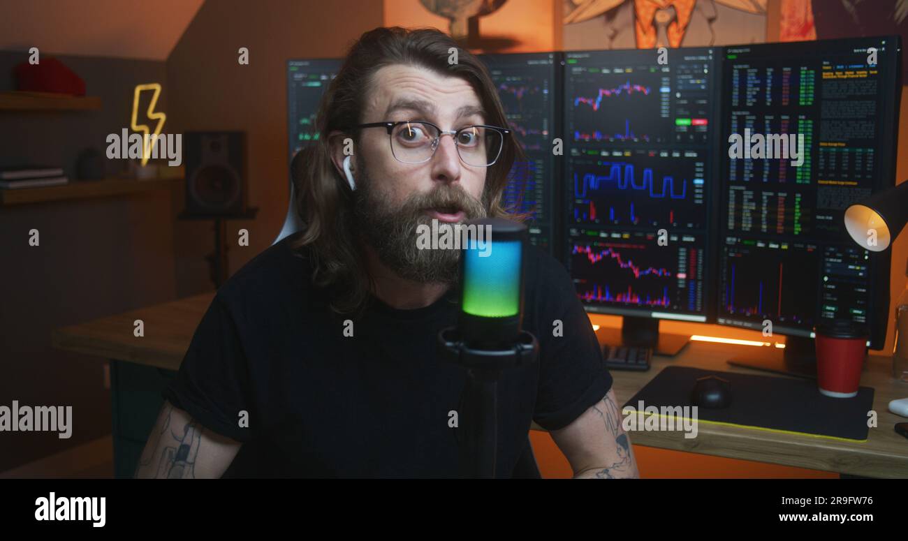 Male stock trader looks at camera and speaks into microphone. Man records educational video training for blog about investment. Multi-monitor computer with real-time stocks, exchange market charts. Stock Photo