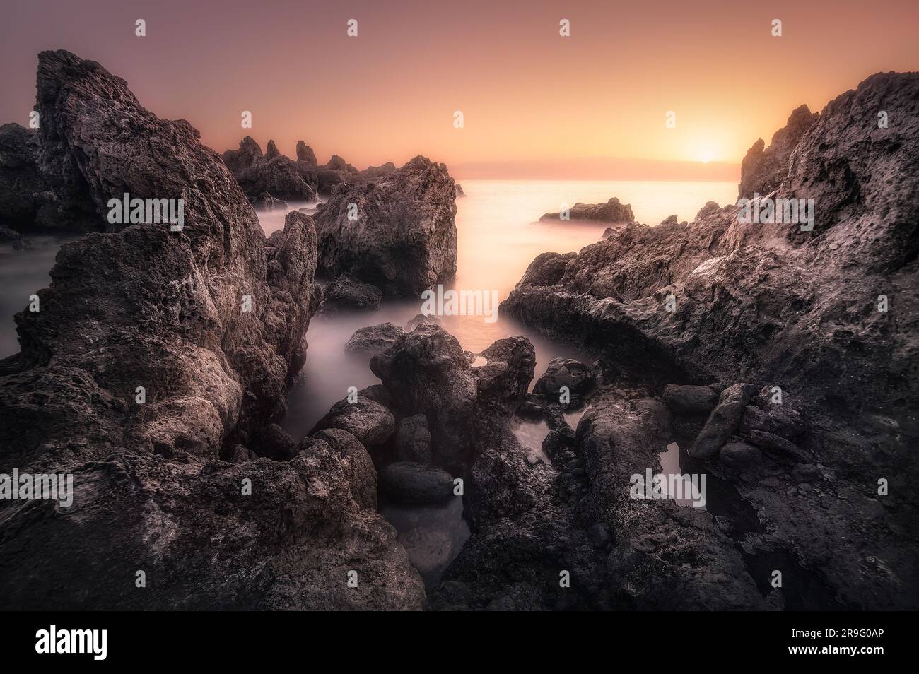 An ocean beach at sunrise, featuring a serene landscape of rocks and gently lapping waves Stock Photo