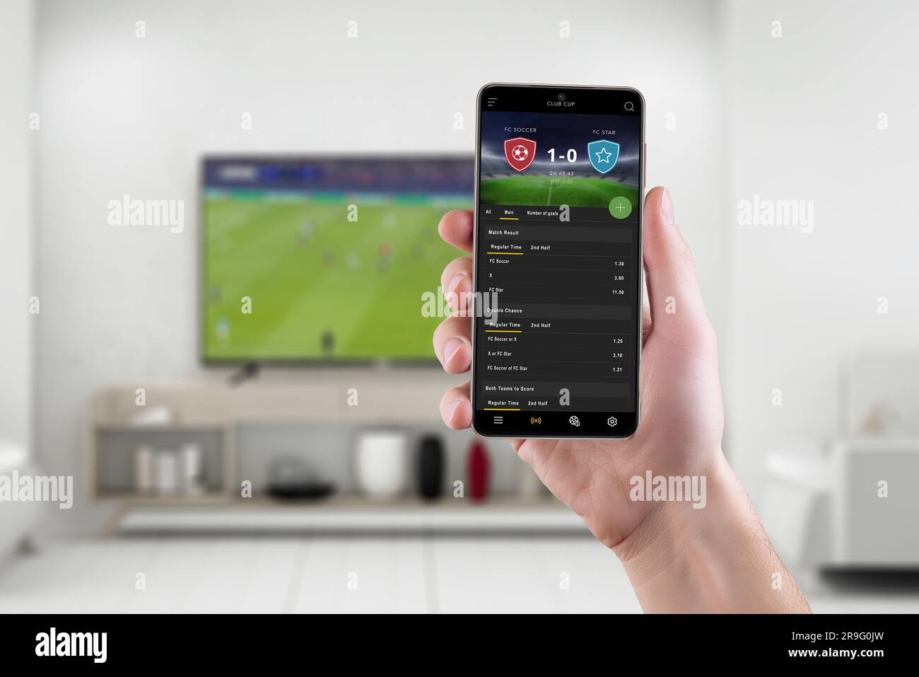 Live soccer betting experience in the comfort of a living room with smart phone. Phone with modern betting app interface in hand. Soccer match broadca Stock Photo