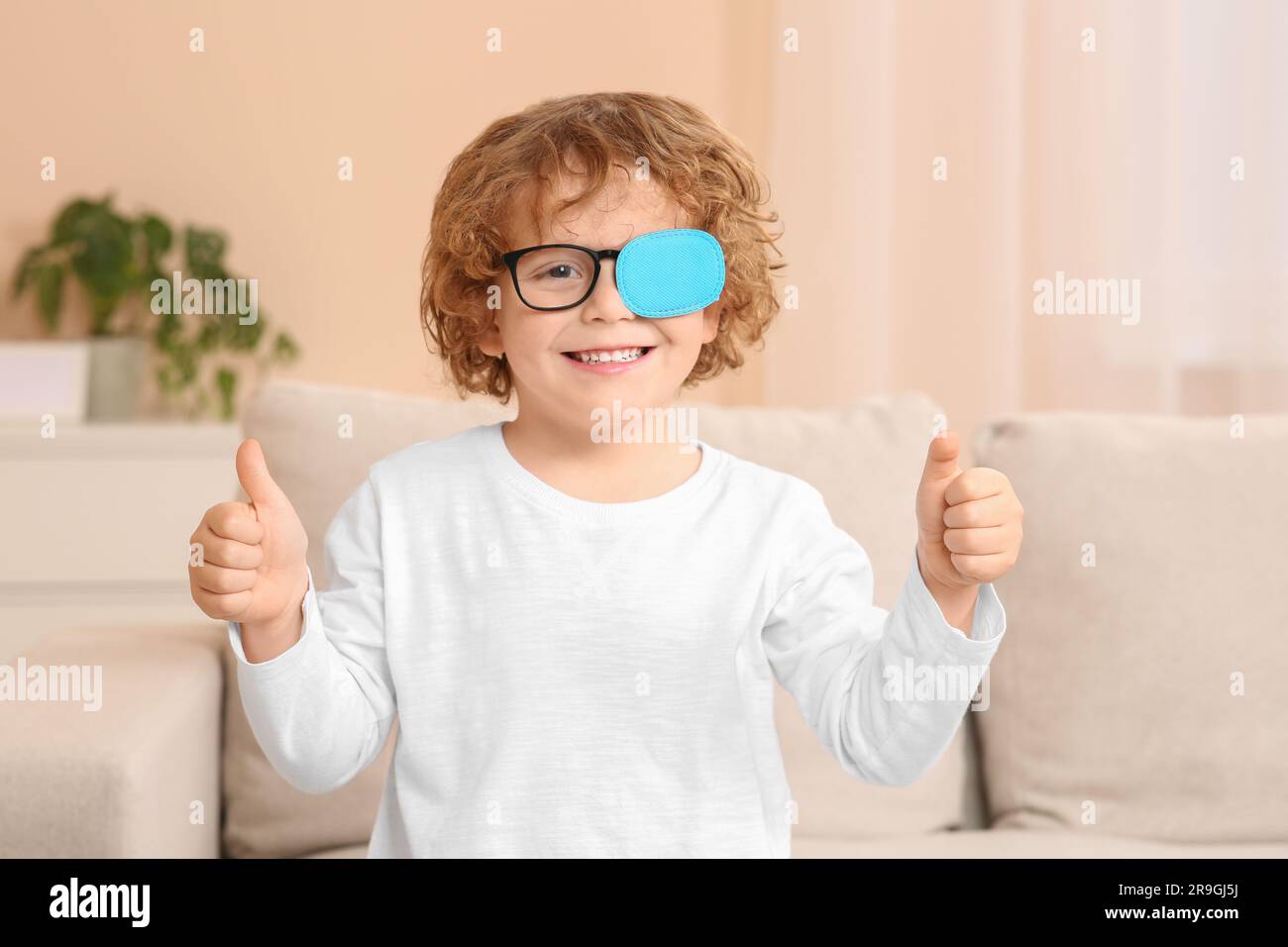 Happy boy with eye patch on glasses showing thumbs up indoors. Strabismus treatment Stock Photo