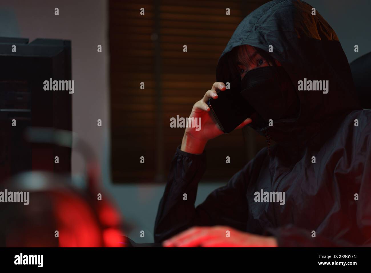 Hacker in hoodie talking on smartphone and typing code to hacking programming system or server. Stock Photo