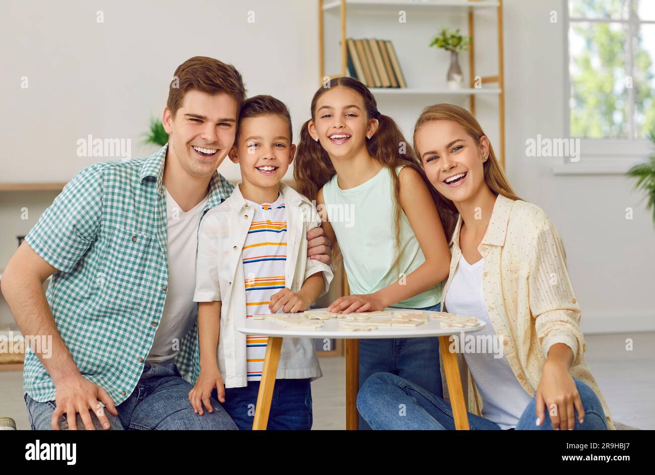 Happy family with two children are playing puzzles at table sitting on floor in living room. Stock Photo