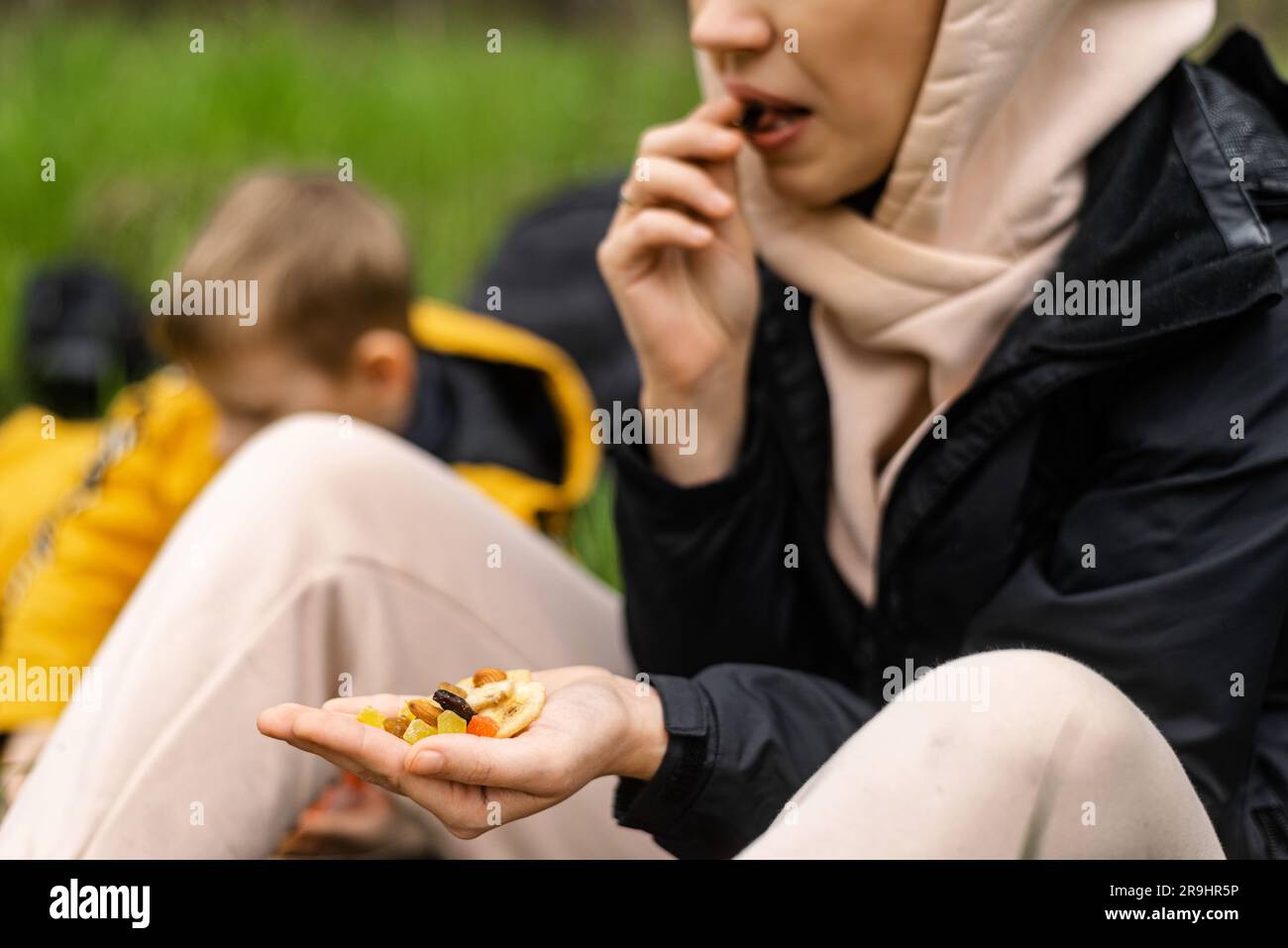 A woman holds various dried fruits and nuts in her hand. Sits on the green grass in the forest. Snack during the hike, walk. Healthy vegetarian food. Stock Photo