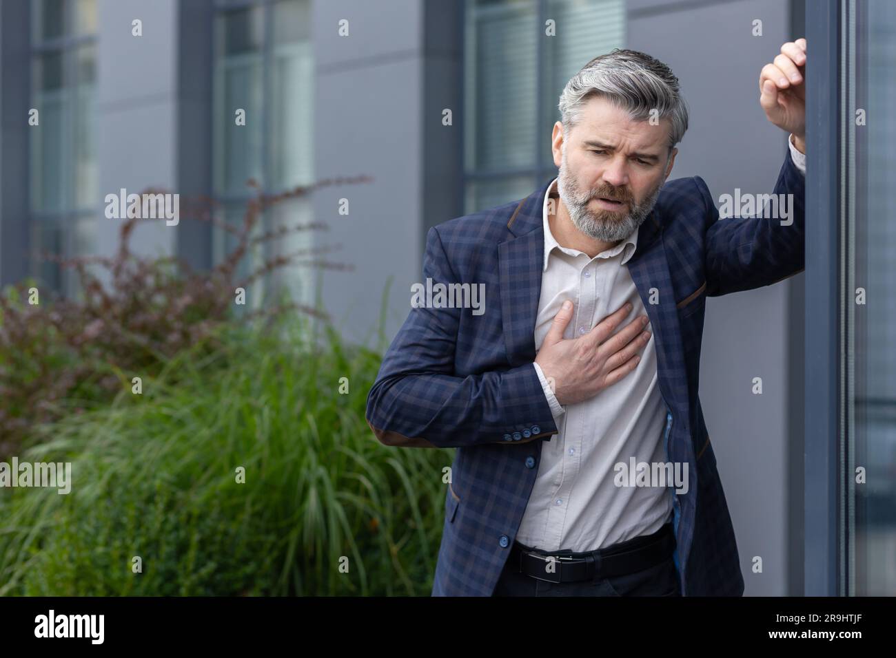 Heart attack and chest pain, mature gray-haired man outside office building has severe pain, businessman boss holding hand to chest and breathing hard from pain. Stock Photo