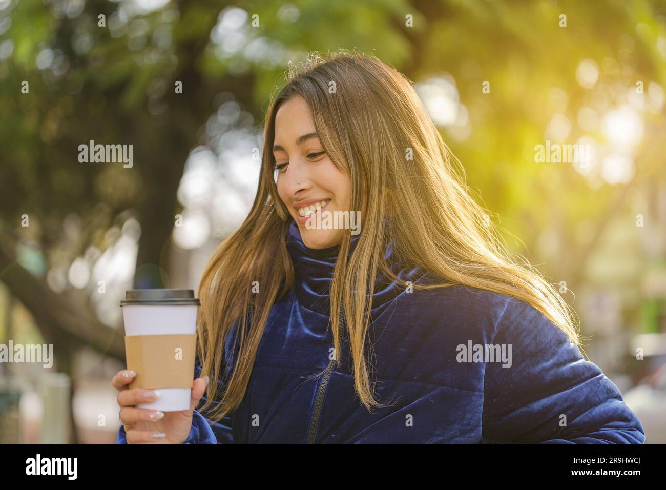 Smiling beautiful latin girl drinking coffee on a public park bench. Stock Photo