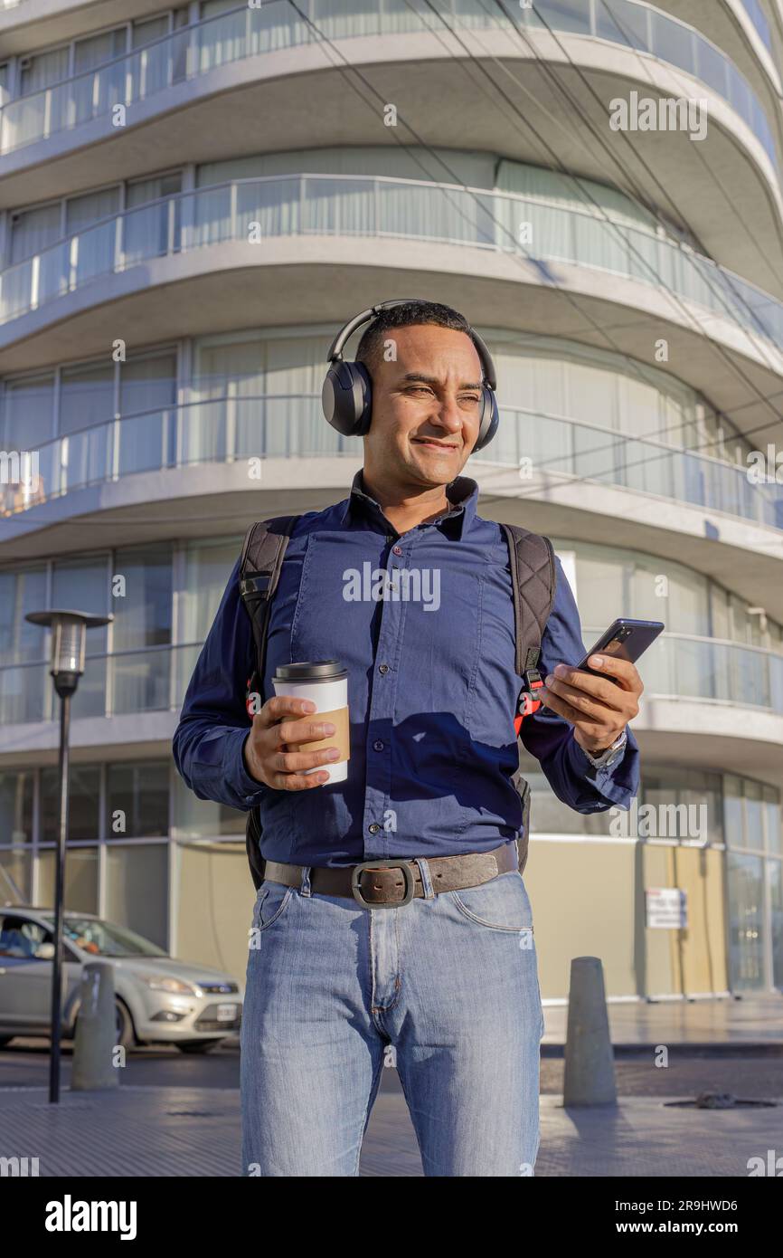 Smiling young latin man with headphones looking at his mobile phone and holding a paper cup with coffee in his hand in a city. Stock Photo