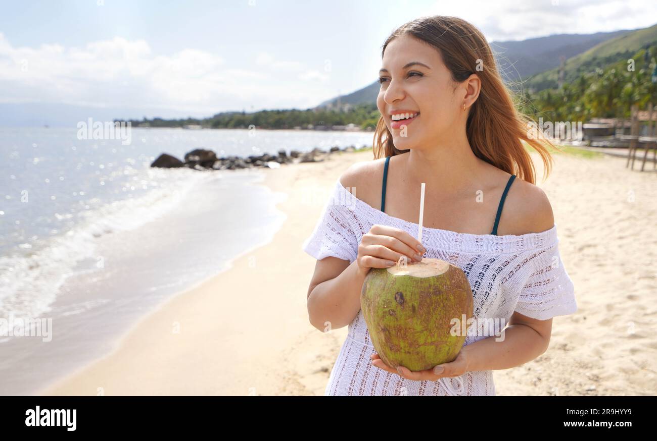 Summer vacation on the beach. Happy relaxed young female tourist drinks green coconut water through a straw. Stock Photo