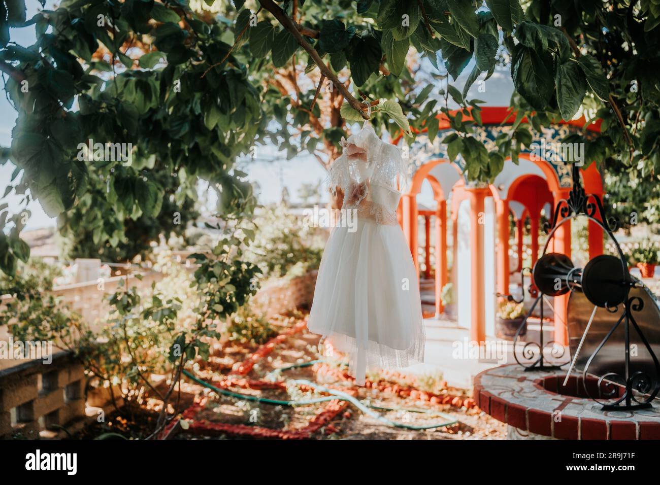 An elegant white infant dress for the baptism ceremony hangs from a tree in an outdoor setting Stock Photo