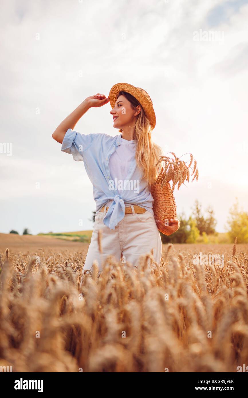 Portrait of young woman walking among wheat in summer field wearing straw hat holding handbag at sunset. Girl relaxing enjoying landscape picking whea Stock Photo