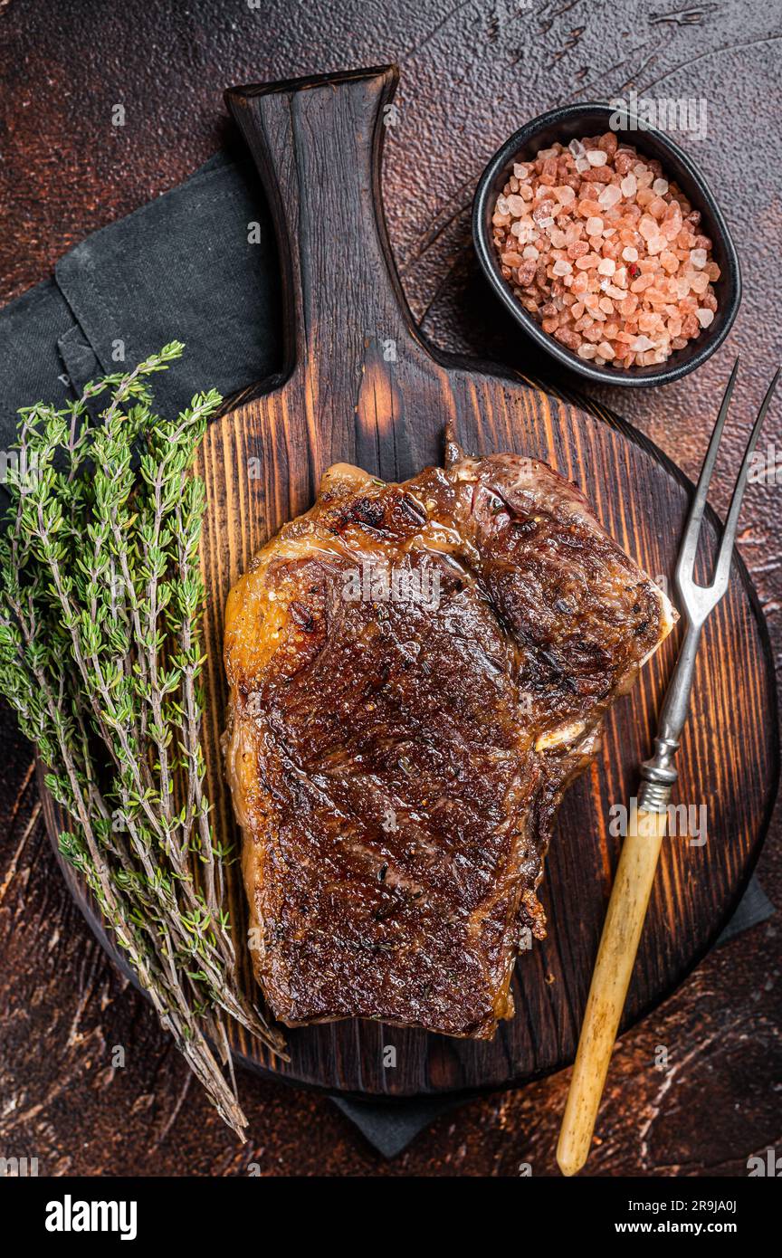 Roast Striploin beef meat steak or club steak on a wooden board with herbs. Dark background. Top view. Stock Photo