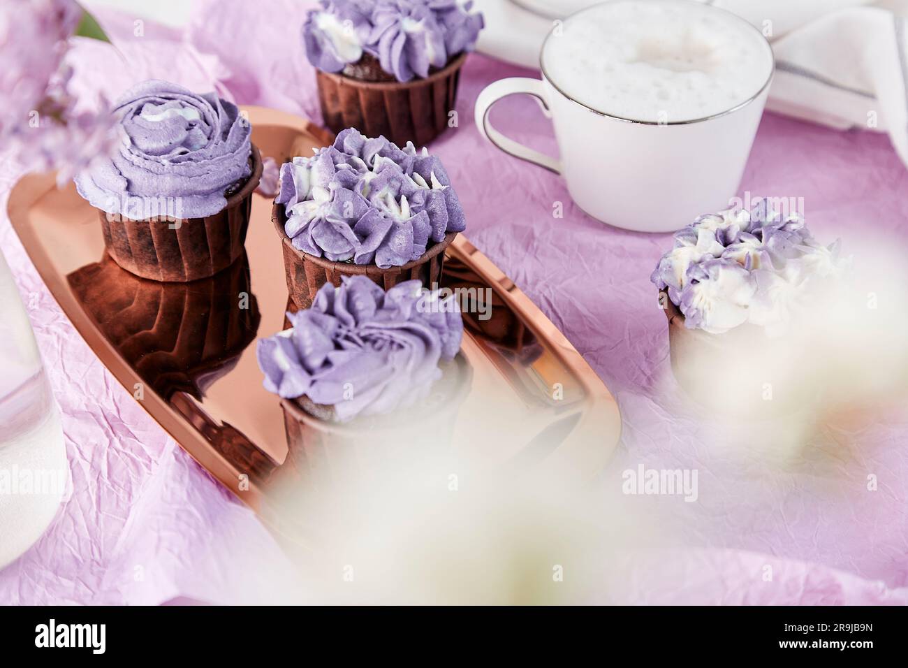 Aesthetics festive sweet table. Purple french cupcakes with cup of coffee among lilac flowers. Feminine lifestyle. Stock Photo