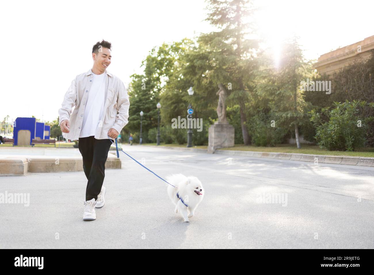 Happy young Asian man in casual clothes with adorable white Pomeranian Spitz dog walking in the park in sunny day Stock Photo