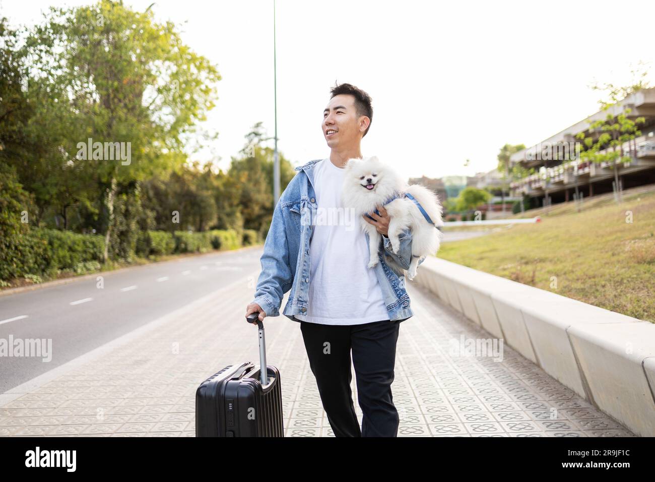 Happy Asian male tourist in casual wear with suitcase and cute Pomeranian Spitz dog smiling and strolling on street Stock Photo