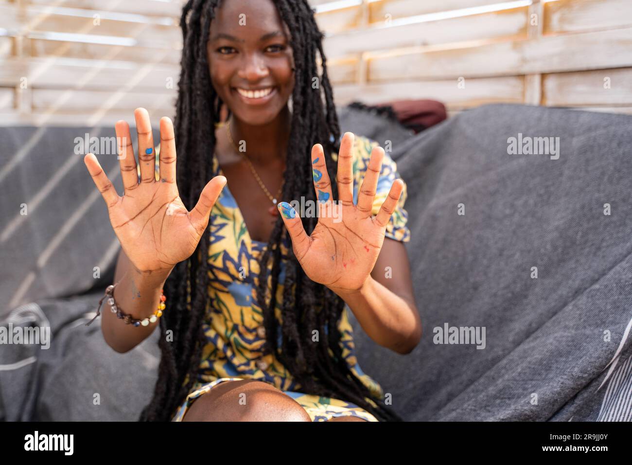 Smiling young African American female in casual clothes with long dreadlocks showing palms with blue paint looking at camera Stock Photo