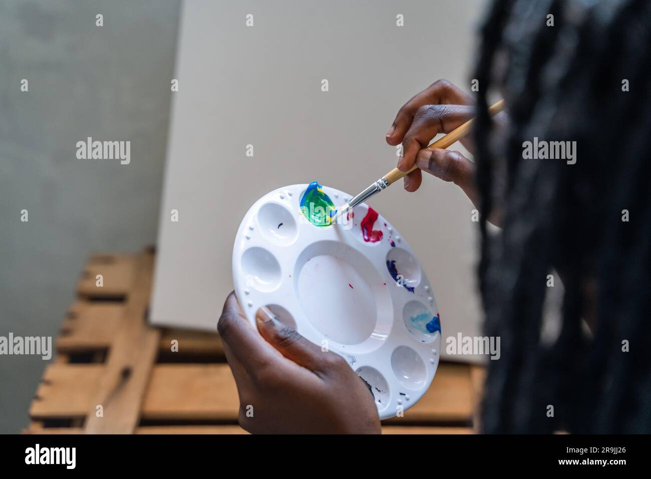 Crop unrecognizable female painter with long deadlocks holding palette while painting in creative studio Stock Photo