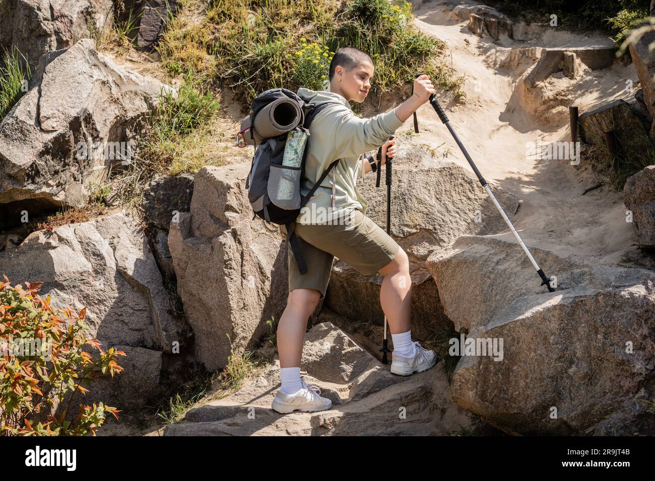 https://c8.alamy.com/comp/2R9JT4B/young-short-haired-female-traveler-in-casual-clothes-with-backpack-holding-trekking-poles-while-standing-near-hill-with-stones-at-background-during-su-2R9JT4B.jpg