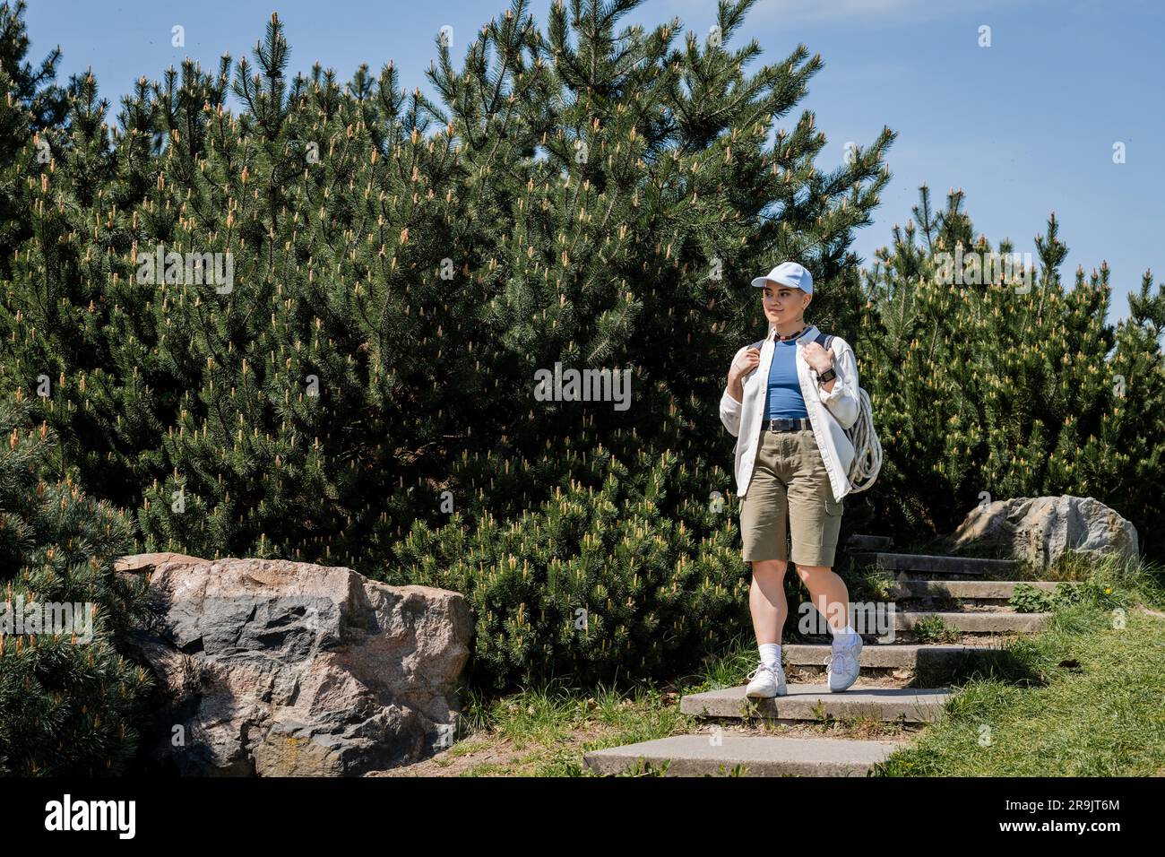 Young female hiker in casual clothes and baseball cap holding backpack while walking down on stairs with nature and blue sky at background, curious hi Stock Photo