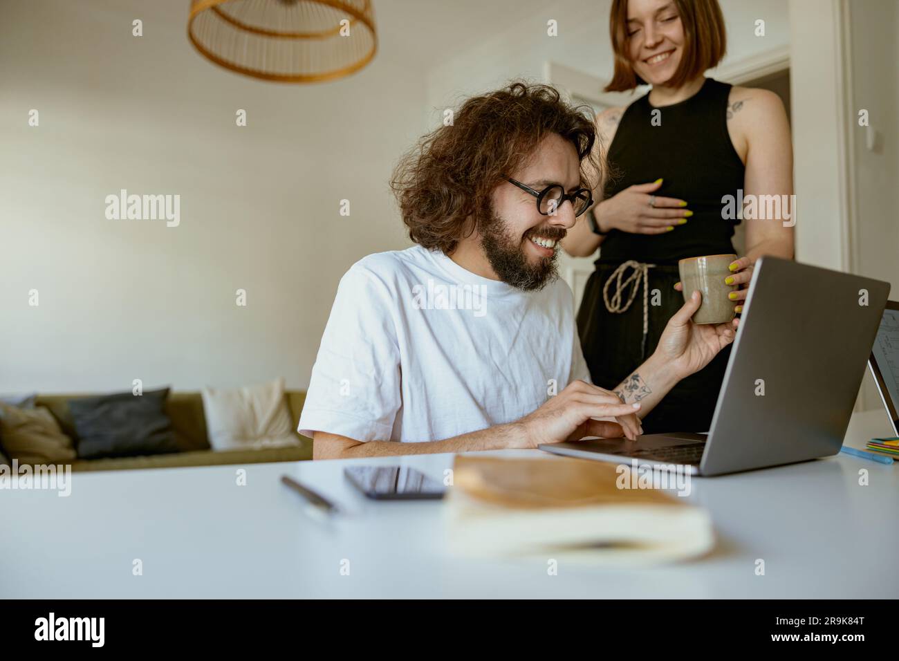 Young smiling man working on laptop from home while girlfriend is brought him coffee Stock Photo