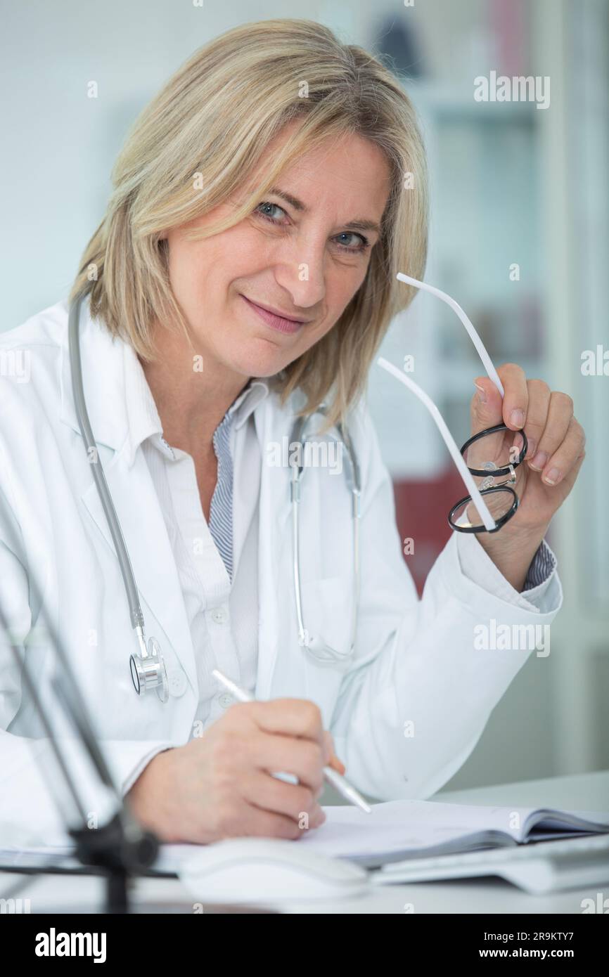 Mature Female Doctor Sat At Her Desk Holding Her Eyeglasses Stock Photo mature-female-doctor-sat-at-her-desk-holding-her-eyeglasses-stock-photo