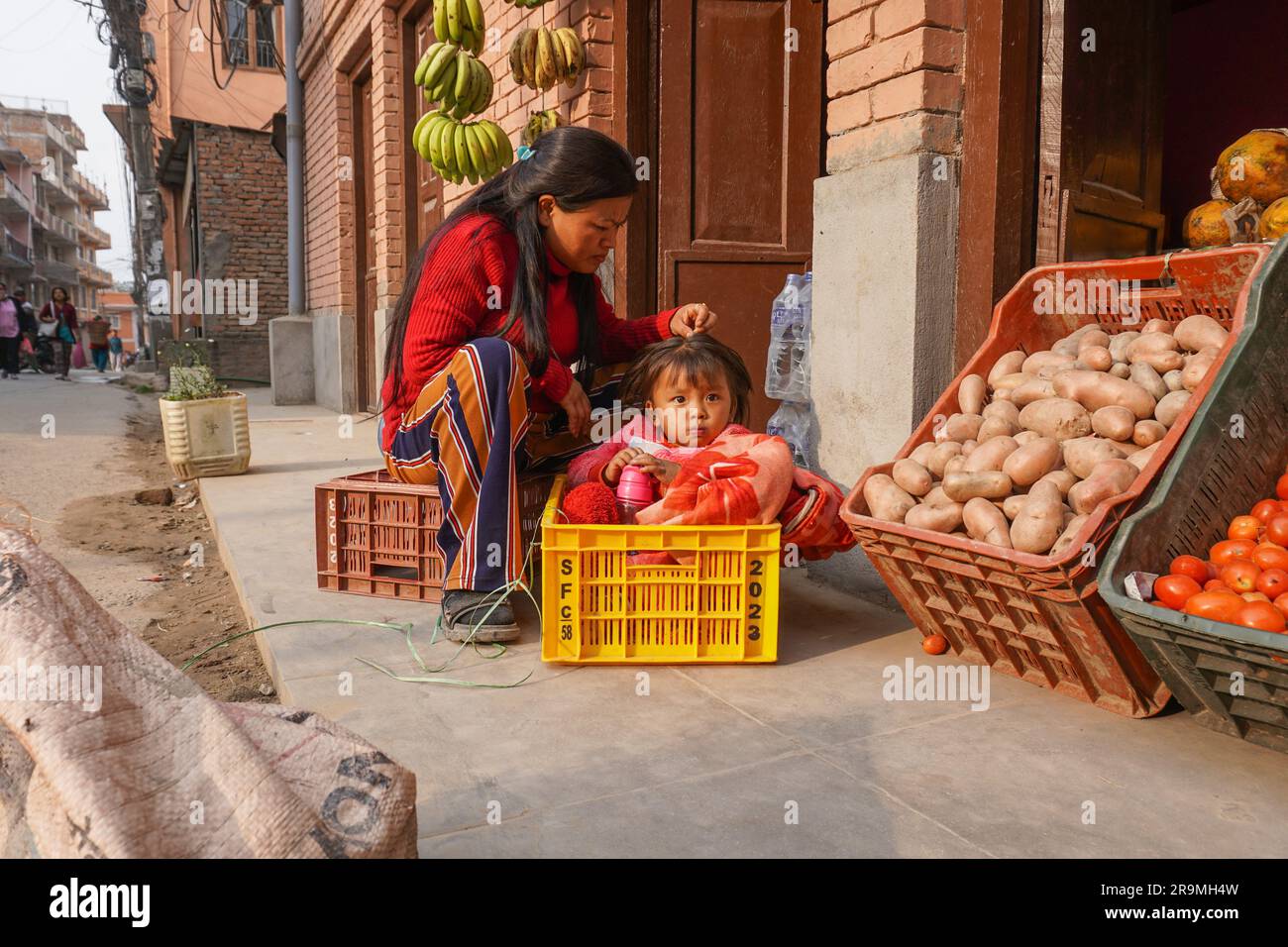 Sita Praja sells fruit in a shop while styling the hair of her 2-year-old daughter, Srishna Praja, in Kirtipur, Kathmandu, Nepal on April 26, 2023. (Sunita Neupane/Global Press Journal) Stock Photo