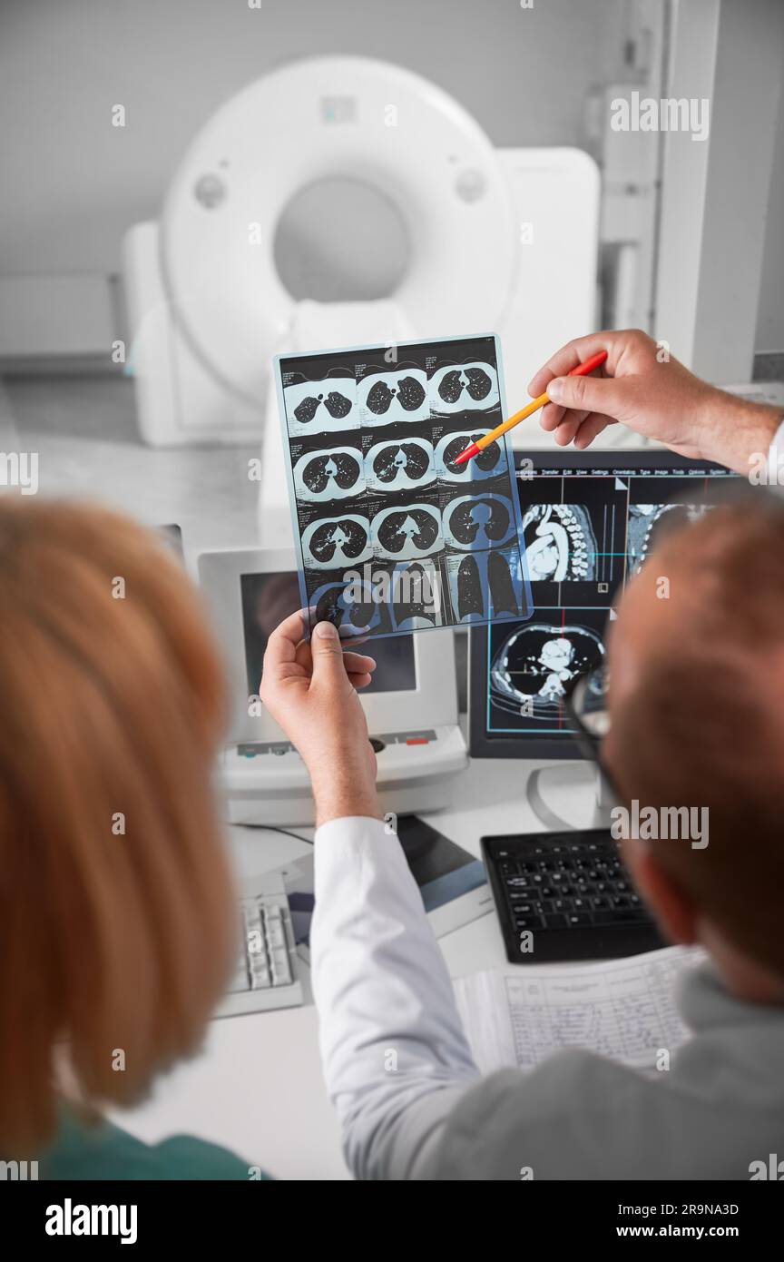 Medical computed tomography or MRI scanner. Back view of doctor and nurse sitting at computer, looking at MRI results. Doctor holding scan, showing, pointing by pen, studying, examining. Stock Photo