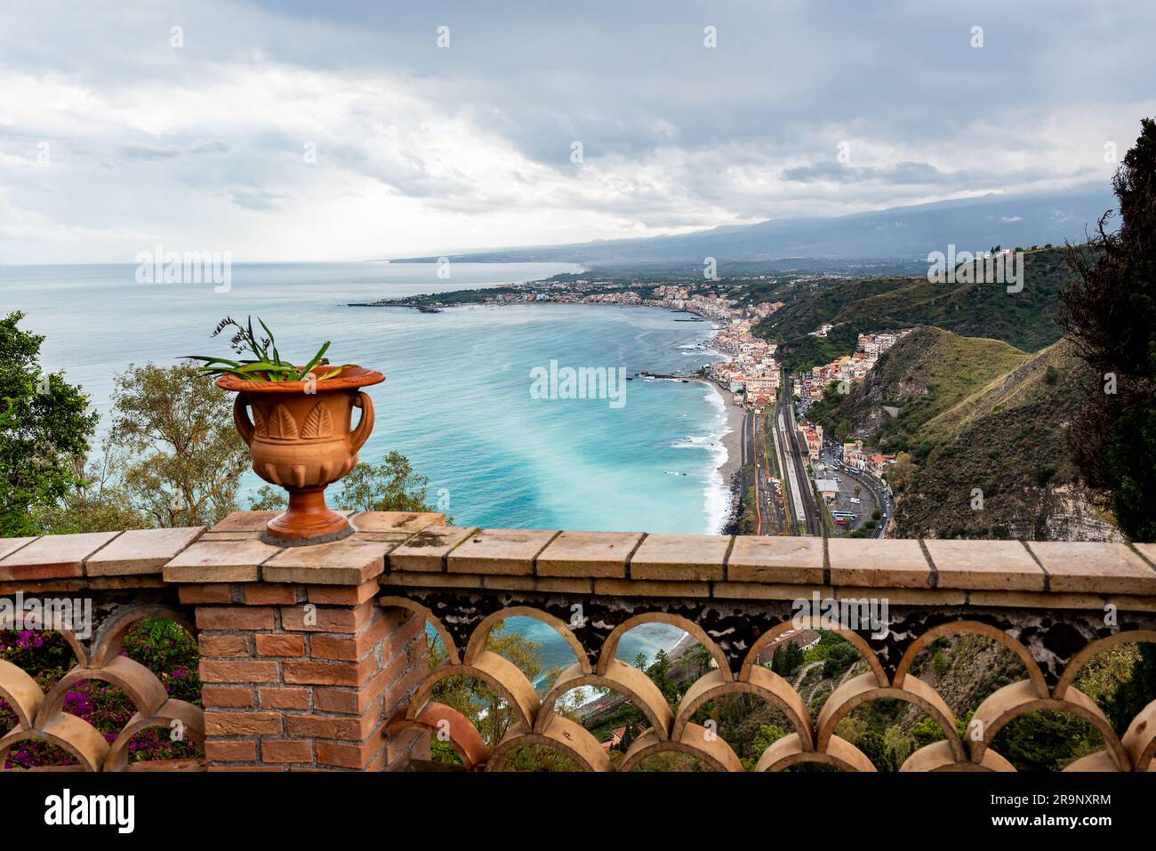 Mediterranean flowering plant in a stone ceramic pot with scenic view on seaside and Mount Etna volcano from public garden Parco Duca di Cesaro to Gia Stock Photo