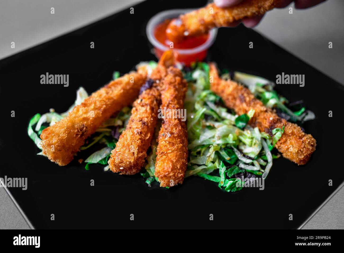 Fried breaded shrimp on vegetable and herb leaf on black plate, closeup. Hand dips shrimp into