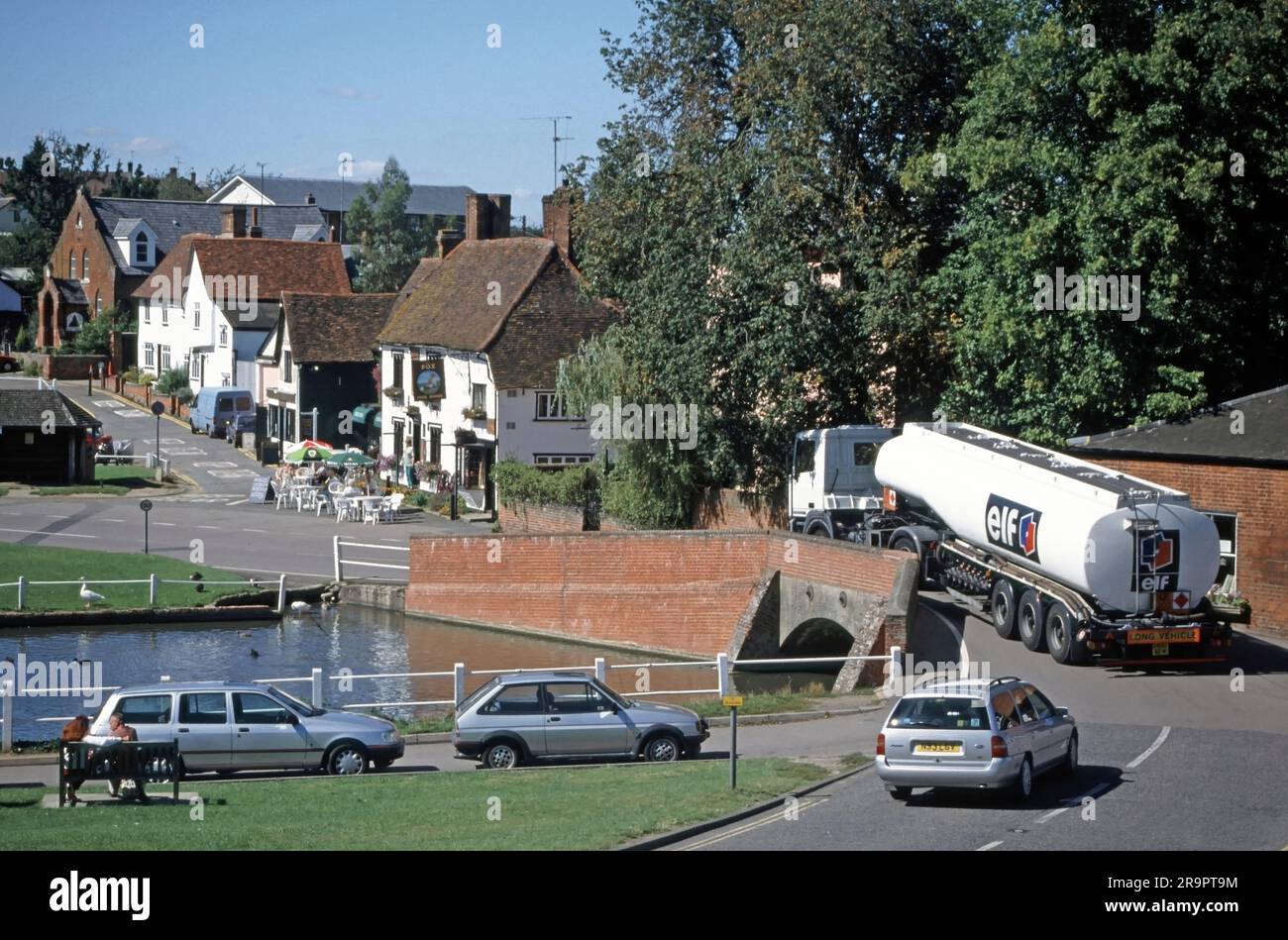 Archival 1994 image of Elf Aquitaine brand and logo on side and back rear end of fuel delivery tanker semi articulated trailer pulled along by hgv lorry truck power unit maneuvering across narrow hump back road bridge in village landscape at Finchingfield in Essex including village pond and car parking in this popular English picture postcard type of visitor attraction in England UK Stock Photo