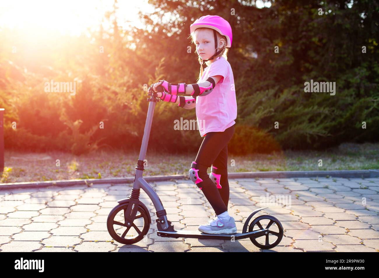A beautiful girl in a pink protective outfit rides a scooter through the streets of the city in summer. Safe cycling and scootering without injury, mo Stock Photo