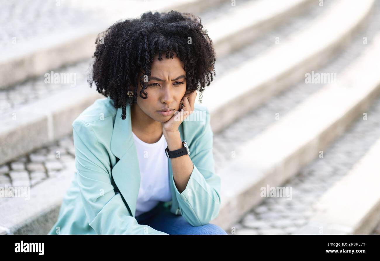 Portrait of frustrated pensive millennial black woman sitting on street Stock Photo