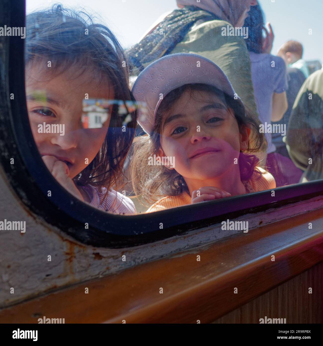 Two very cute endearing cheeky little girls look into the window of a passenger ferry with childhood curiosity in Istanbul Turkey Stock Photo