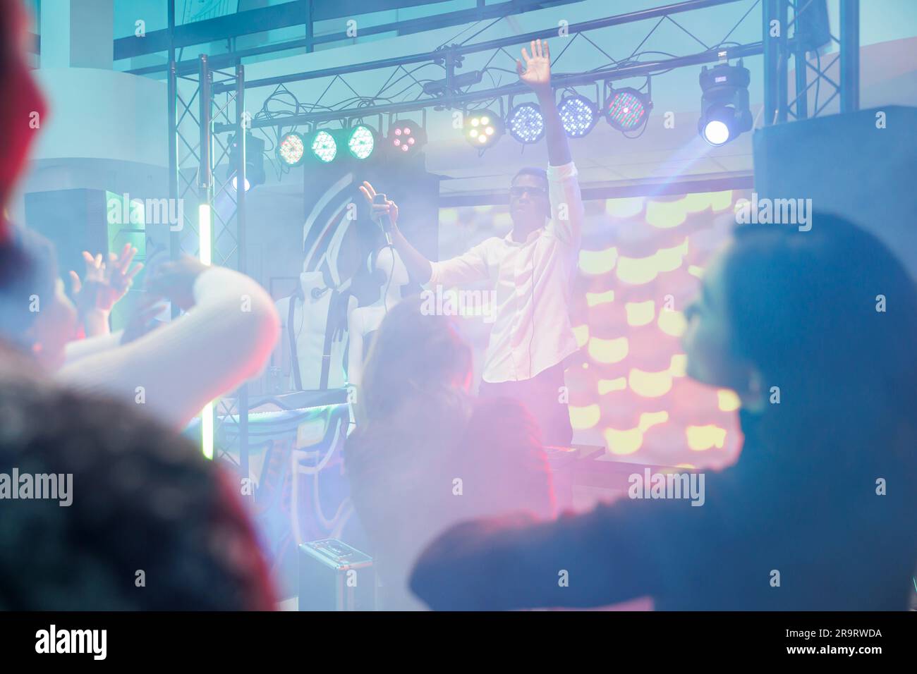 Musician with microphone raising hand while entertaining crowd from stage in nightclub. Dj singing and playing at electronic music show and discotheque party in club with spotlights Stock Photo