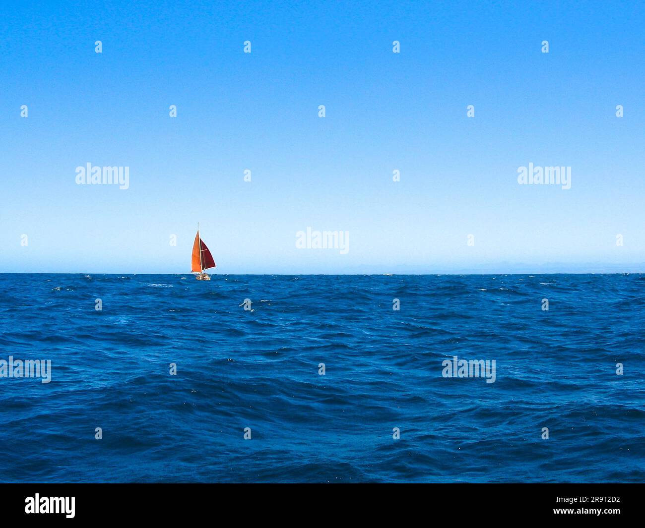 Small sailing boat with red sails on crisp blue horizon, blue ocean and perfect trade wind sea voyage. Stock Photo