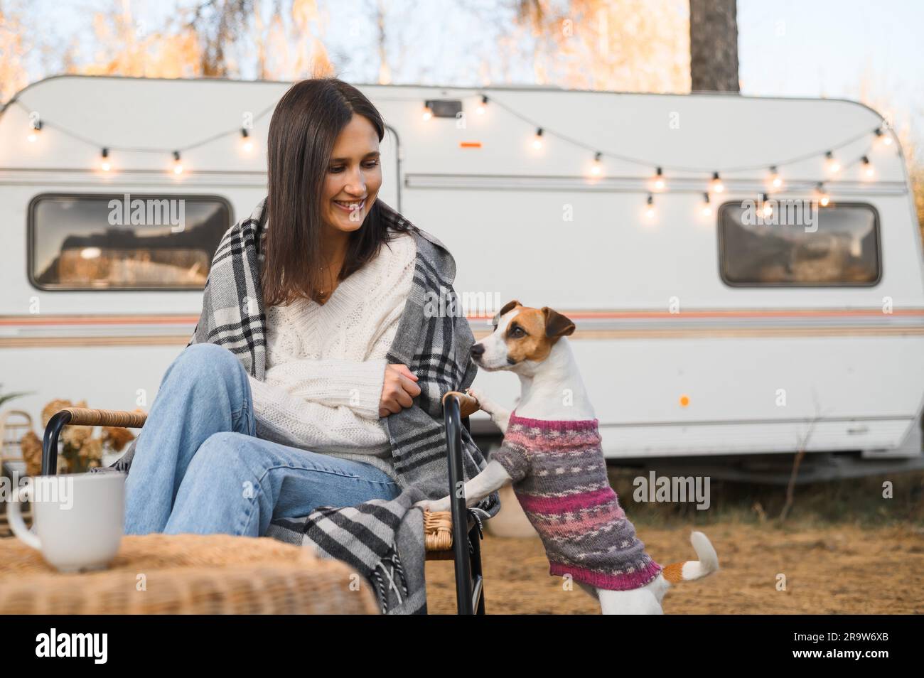 Caucasian woman sitting in a wicker chair wrapped in a blanket with a dog in the yard near the trailer in autumn.  Stock Photo