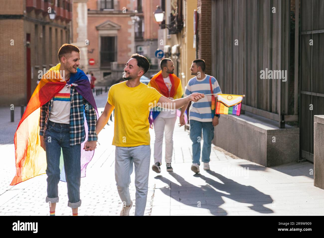 In a sun-kissed city, two gay couples radiate pure happiness, proudly adorned with LGBT symbols while holding hands, immersed in a lively sunset walk. Stock Photo