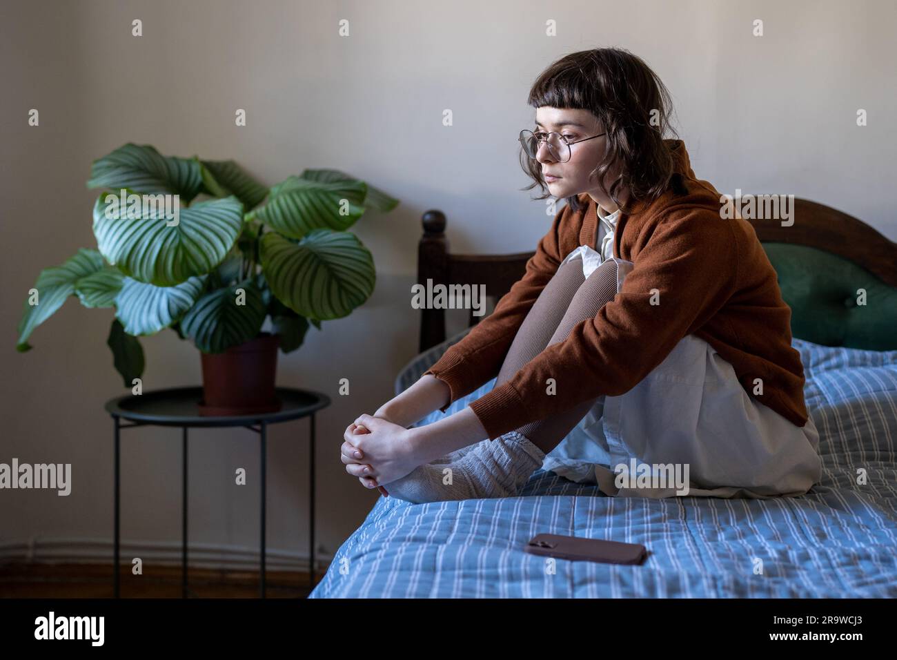 Pensive sad young woman sits on bed, hugs knees next to mobile phone waiting for call, message, sms Stock Photo