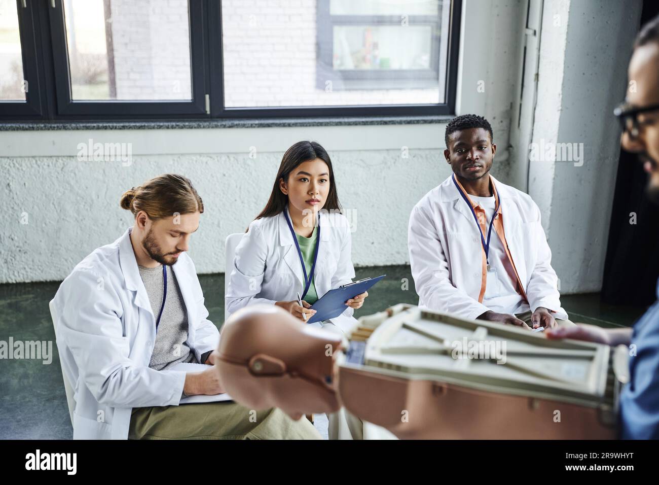 medical training, diverse group of multiethnic students in white coats looking at paramedic standing with CPR manikin on blurred foreground, acquiring Stock Photo
