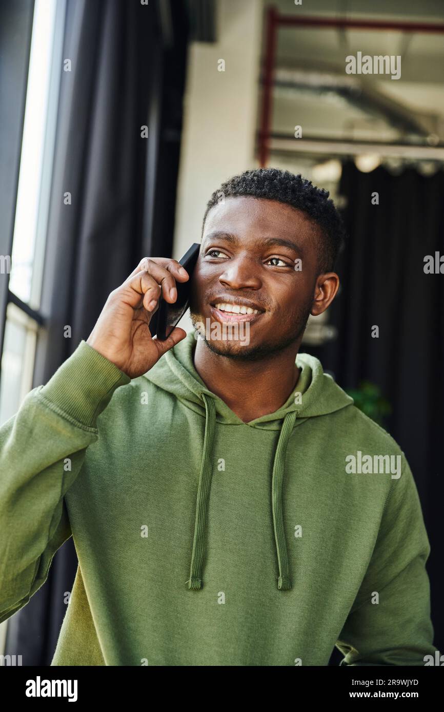 optimistic african american man in green hoodie talking on mobile phone and looking away in contemporary office, successful entrepreneur discussing bu Stock Photo