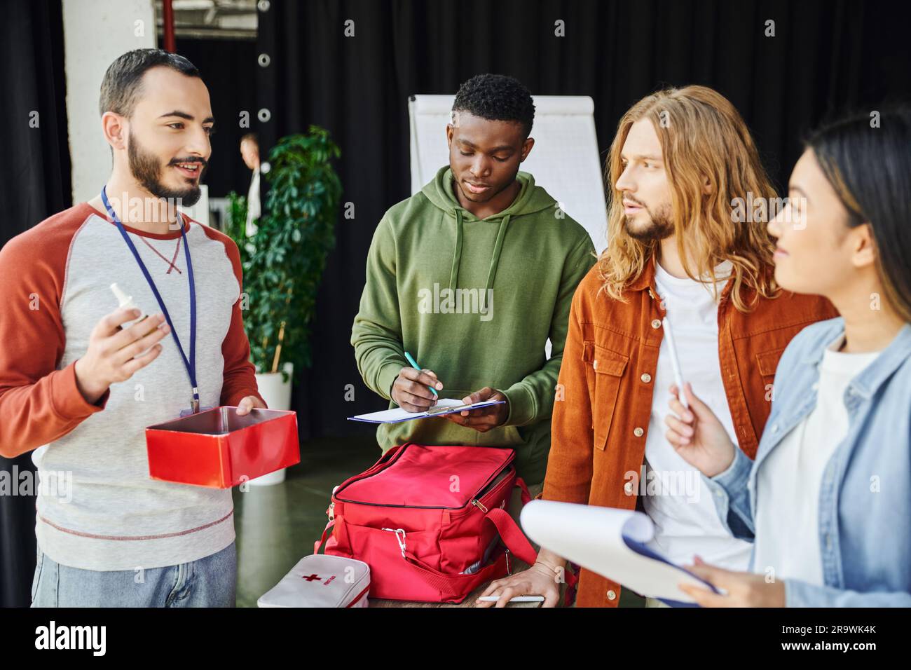 bearded paramedic talking and showing nasal spray and first aid kit to multicultural team of young participants in first aid seminar, safety and emerg Stock Photo