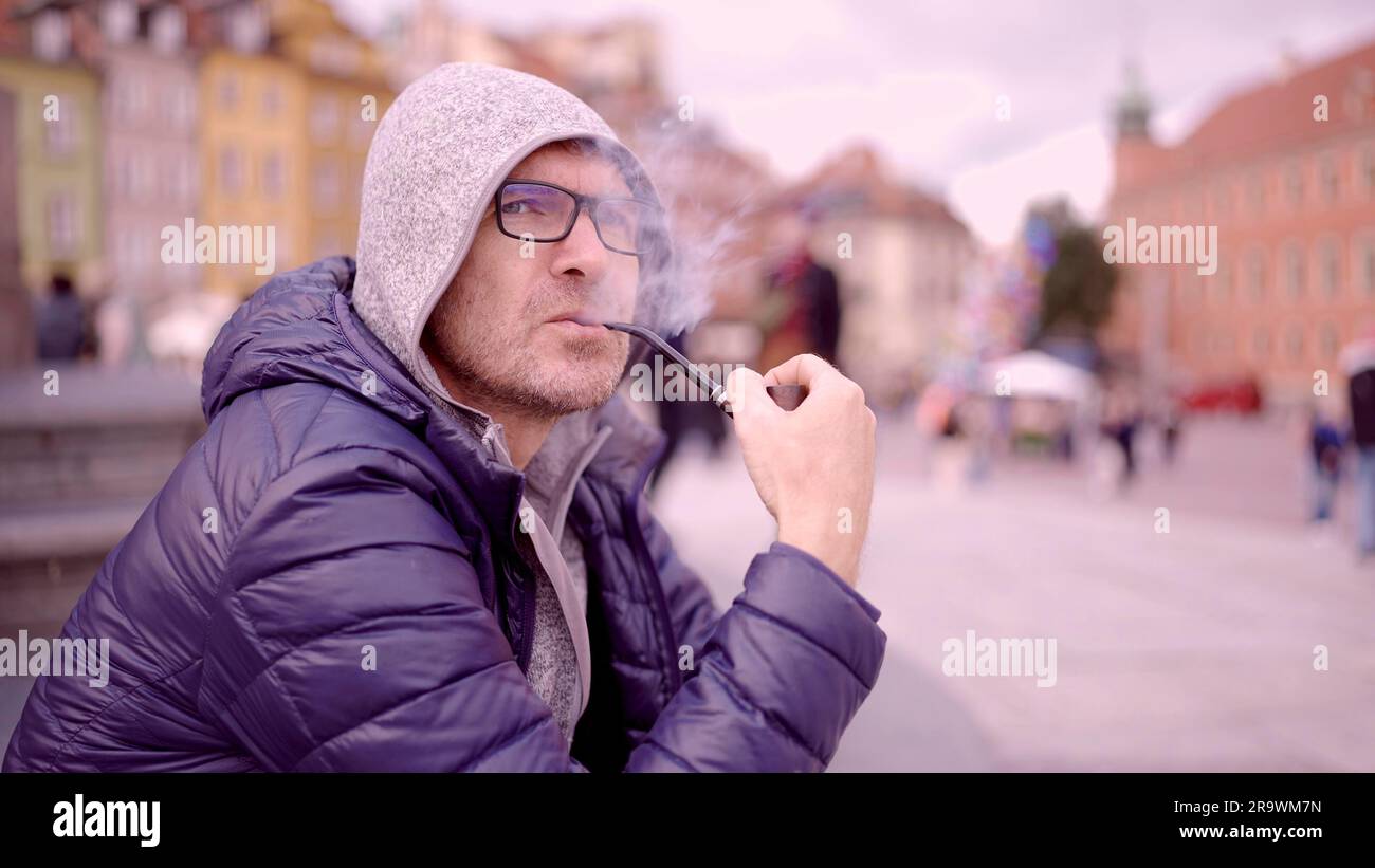 Adult man with glasses sitting on square and smoking a tobacco pipe releasing smoke in the Palace Square, Warsaw Old Town Stock Photo