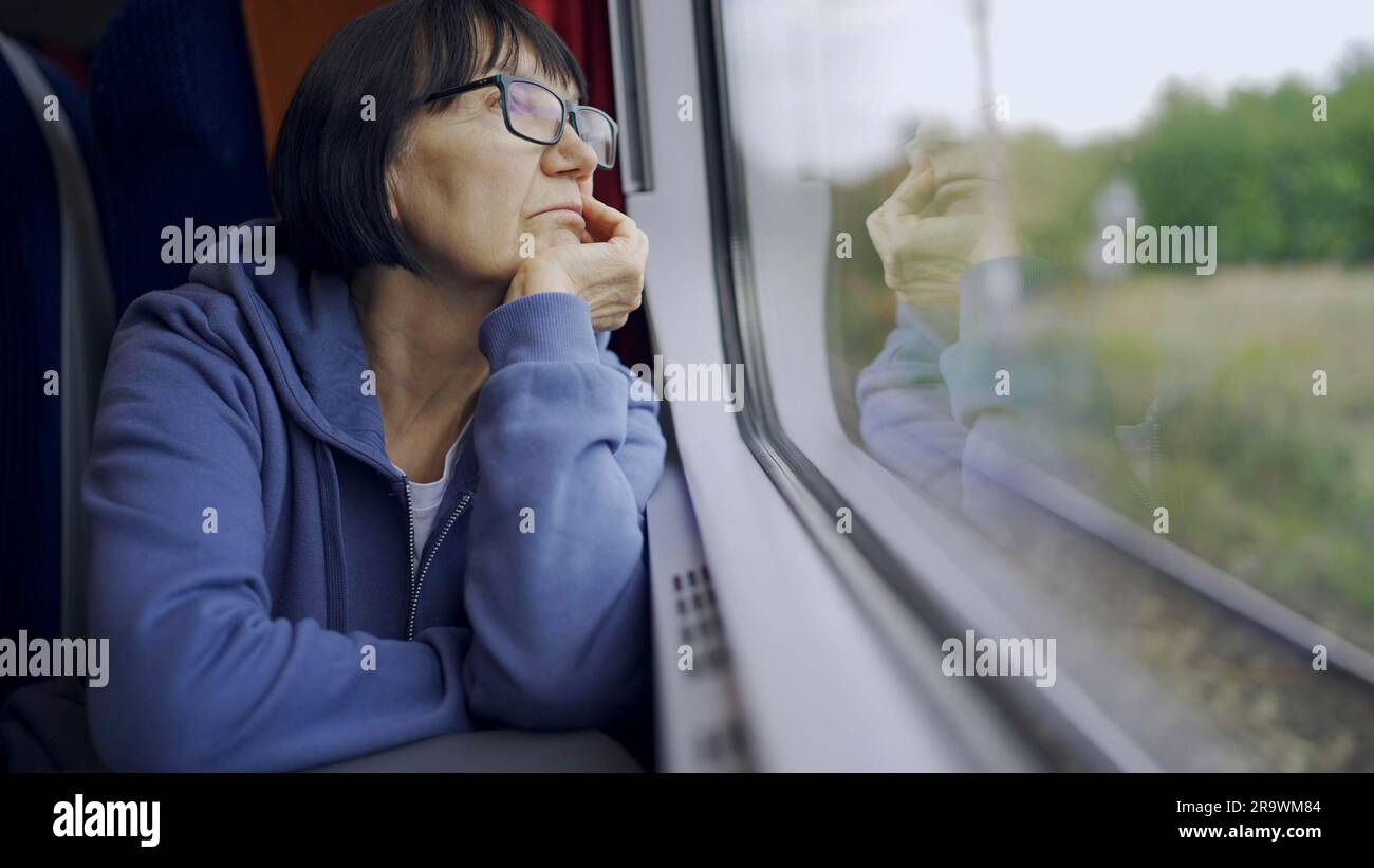 Elderly lady in glasses travels in train and looking out the window reflected in the glass Stock Photo