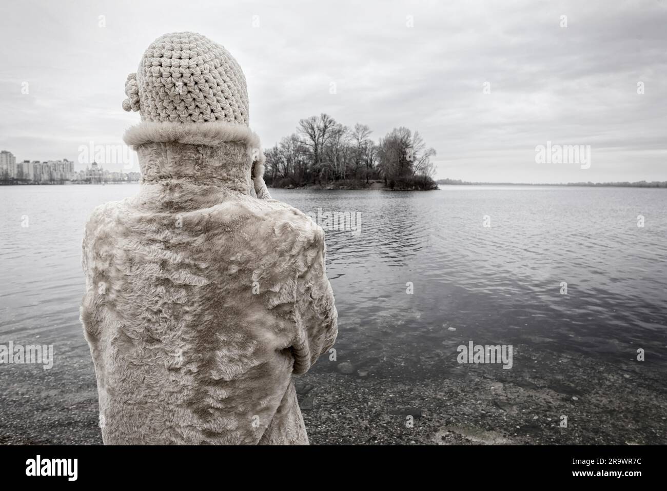 A woman with a wool cap and a fur coat is looking at the river during a cold and sad gray winter morning Stock Photo