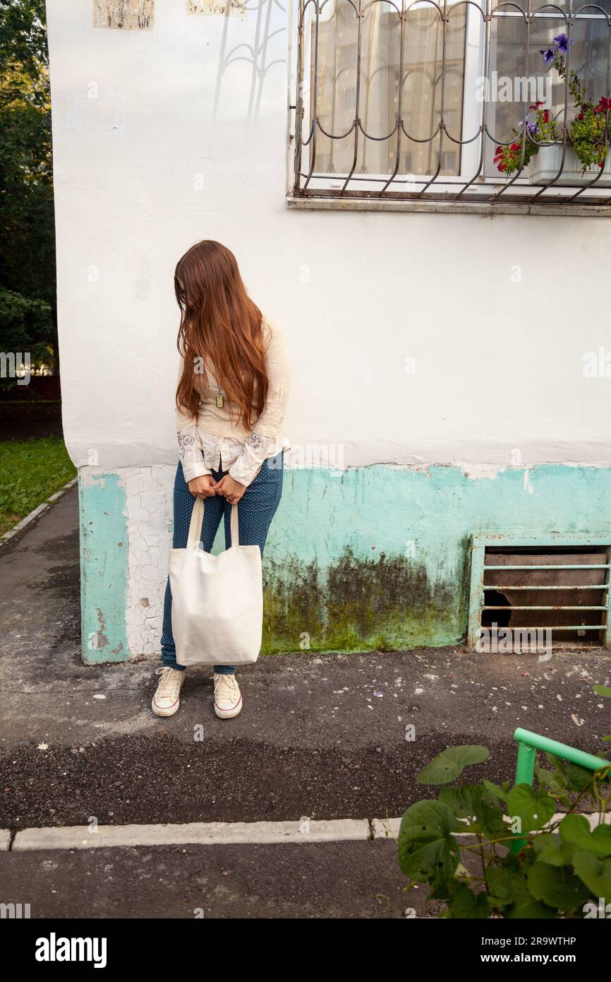 Young long-haired woman with a bag standing on a street and waiting. Pensive, worried Caucasian lady with brown hair looking down - Stock Image