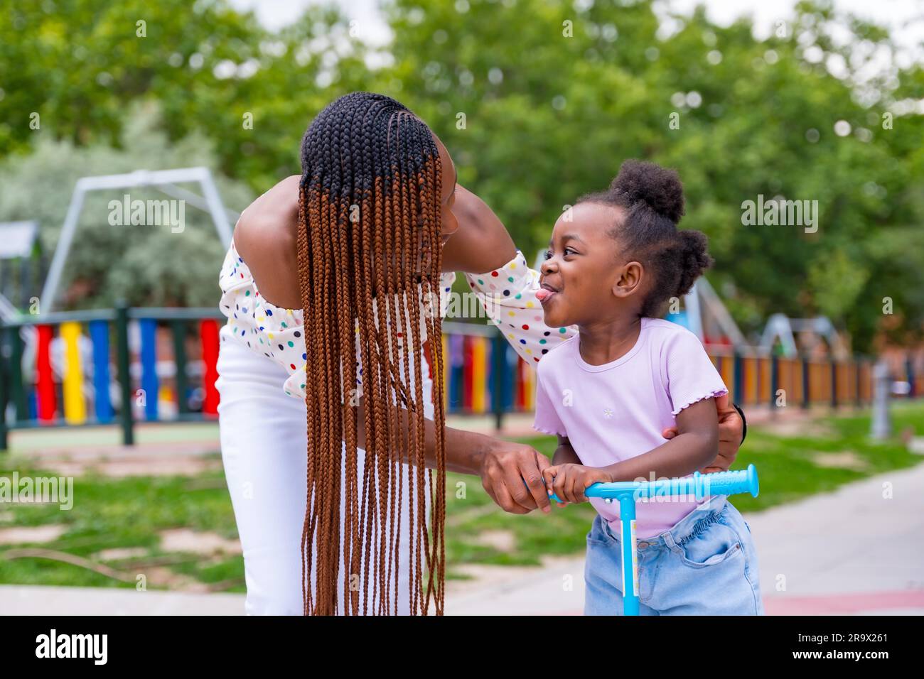 Black African ethnicity mother having fun with her daughter in playground walking with the skateboard and sticking out her tongue Stock Photo